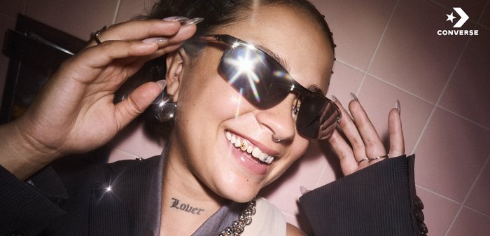 Zonnebril met donkere lenzen en een metalen frame, met nadruk op reflectie en glans. Persoon toont een tandaccessoire en tattoo.