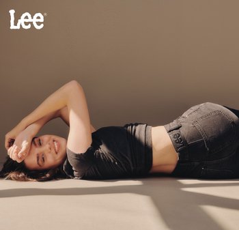 Jeans neri in denim con dettagli di cucitura visibili e una toppa con logo. Il modello è sdraiato su una superficie, indossando un top nero aderente. La luce naturale mette in risalto le texture.