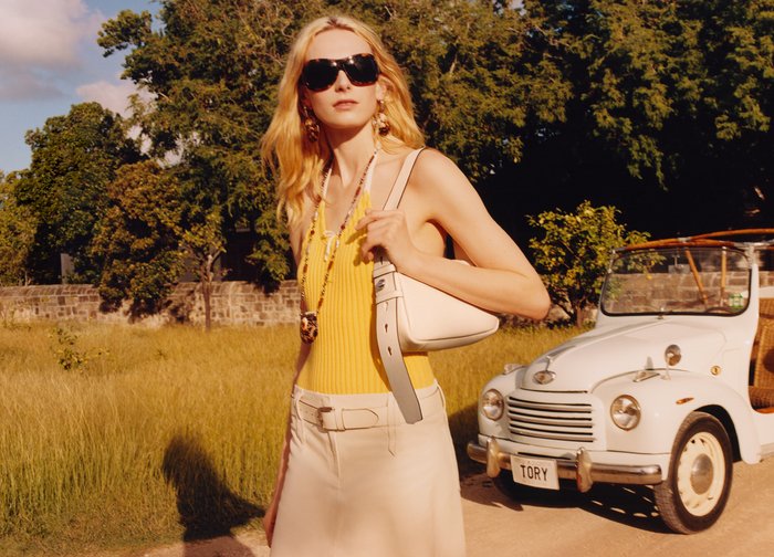 Femme blonde portant des lunettes de soleil, un haut jaune et un pantalon blanc, debout à l'extérieur près d'une voiture blanche vintage avec une plaque d'immatriculation "TORY".