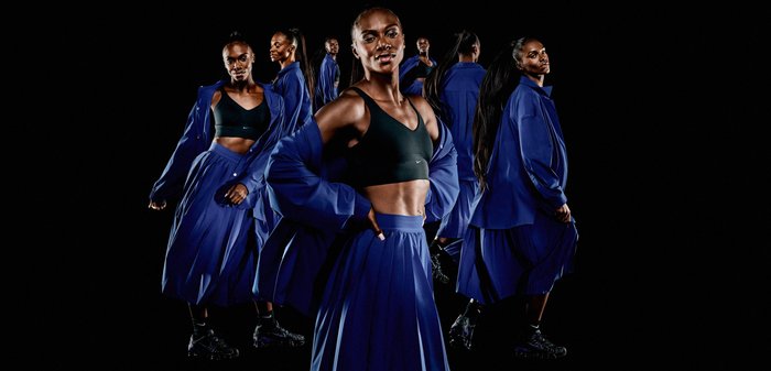 Model wears a black sports bra and a long blue skirt, layered with a loose blue jacket. Features smooth fabric and simple design.