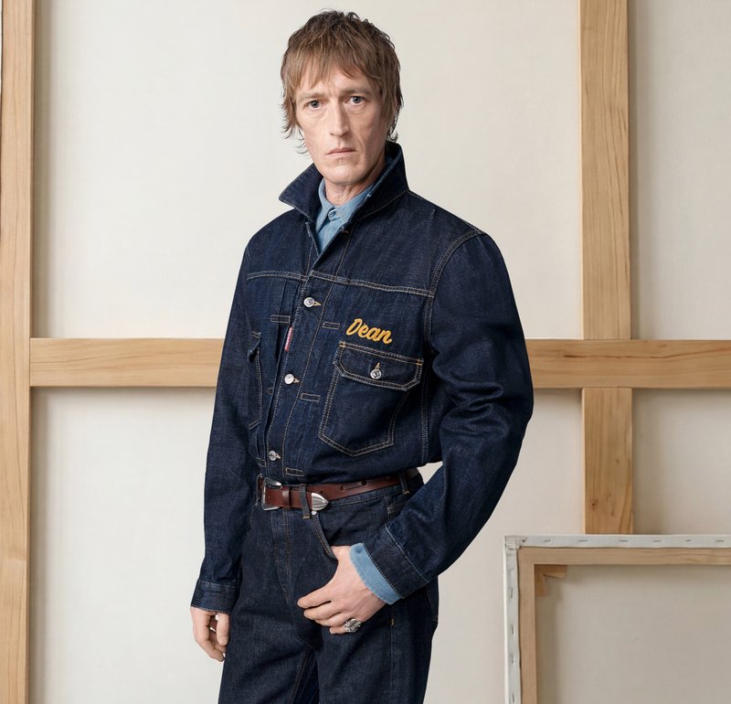 Homme debout en veste en jean et jeans à côté d'un mannequin, avec des rouleaux de tissu et des étagères en bois dans un atelier.