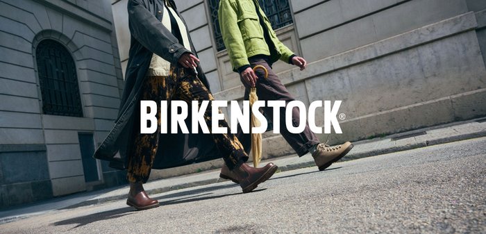 Braune Leder-Ankle-Boots und grüne Jacke, kombiniert mit gemusterten Hosen, während in einem städtischen Umfeld mit einem sichtbar in der Hand gehaltenen Regenschirm gegangen wird.