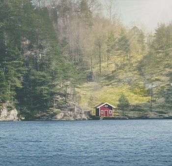 Maison en bois rouge avec des bordures blanches, de forme rectangulaire, toit incliné, située près d'un plan d'eau avec en toile de fond des arbres et une colline herbeuse.