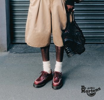 Persona che indossa pantaloncini beige, calzini bianchi, scarpe in pelle color bordeaux, che tiene una borsa nera a rete, in piedi su un marciapiede grigio davanti a una serranda metallica.