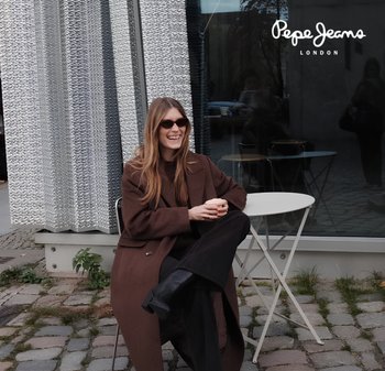Brown long coat, black pants, and knee-high boots. Gray background with textured panels. Round white table with black stools nearby.