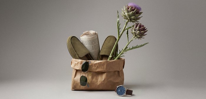 Smeđa papirnata vrećica sadrži smotani novine, maslinaste papuče, sunčane naočale i dva ljubičasta cvijeta čička; ručni sat s plavim brojčanikom leži u blizini.