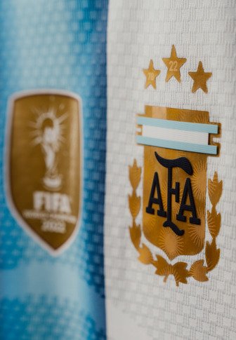 Badge de l'équipe nationale de football d'Argentine avec trois étoiles d'or au-dessus et un emblème flou de la Coupe du Monde de la FIFA 2022 en arrière-plan.