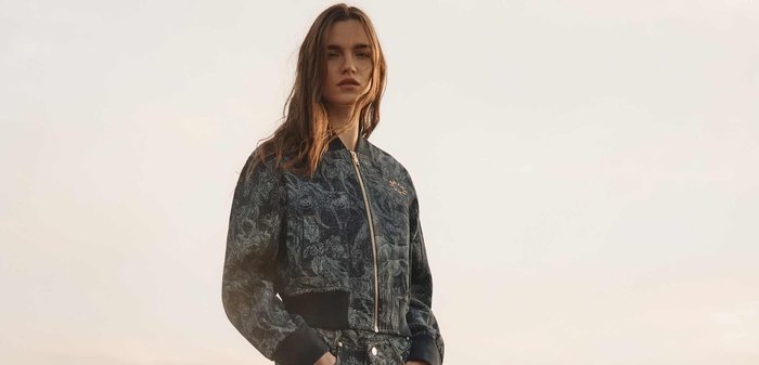 Jeune femme debout dans un paysage désertique, portant une veste à motif sombre et un pantalon large assorti, les mains dans les poches.