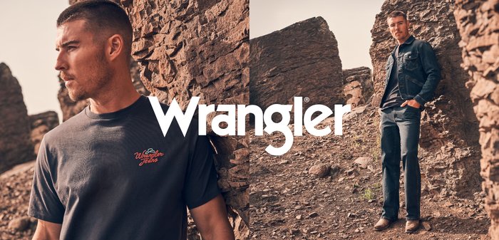 Marinblå t-shirt med röd "Wrangler Jeans"-logotyp, kombinerad med en denimjacka och mörka jeans, placerad mot en bergig terräng.