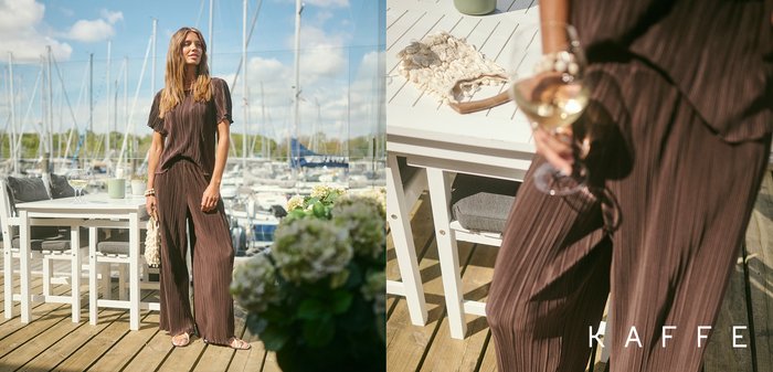 Femme portant des lunettes de soleil, souriante et portant un toast avec du vin blanc à l’extérieur avec d’autres personnes lors d’une journée ensoleillée près d’une marina ou d’un bord de l’eau.