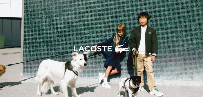 Dvě děti drží vodítka dvou psů. Chlapec má na sobě černou bundu přes bílou košili a dívka má modré šaty. V pozadí jsou světlé dlaždice.