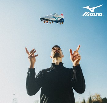 Botas de fútbol Mizuno con una parte superior blanca, acentos azules y detalles rojos, flotando sobre una persona con chaqueta negra contra un cielo despejado.