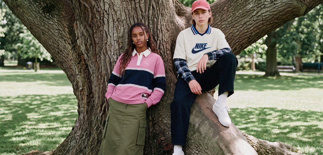 Dos jóvenes vestidos con ropa deportiva casual están de pie y sentados sobre un gran tronco de árbol en un parque soleado.