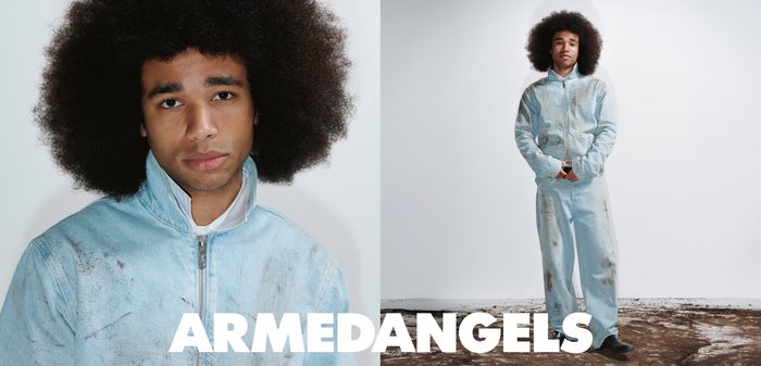 Giovane con un ampio afro che indossa una tuta azzurra sporca, in piedi su terreno contro una parete bianca, testo "ARMEDANGELS" in basso.