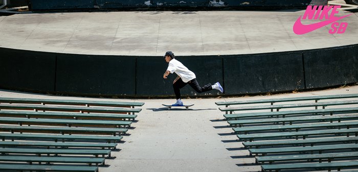 Un skateboarder in t-shirt bianco e pantaloni neri usa uno skateboard su una superficie di cemento, con panchine verdi disposte in modo circolare.