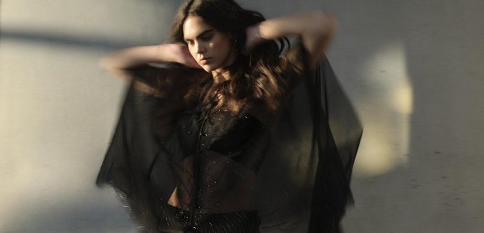 Indumento in tessuto nero trasparente con una vestibilità ampia, caratterizzato da decorazioni. Il modello mette in mostra texture sovrapposte e movimento. Una luce soffusa evidenzia i dettagli.
