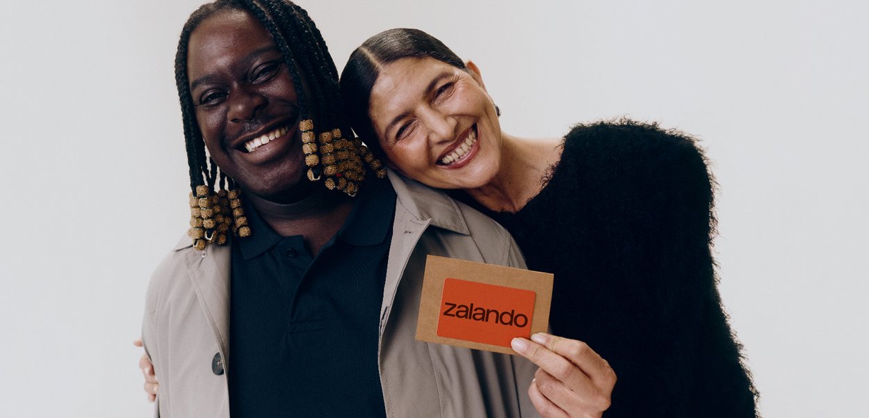 Δύο άτομα που χαμογελούν, ο ένας με ανοιχτόχρωμο παλτό τύπου trench, ο άλλος με υφασμάτινη μαύρη μπλούζα, κρατώντας μια κάρτα Zalando.