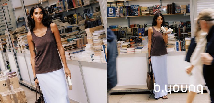 Frau mit langen dunklen Haaren hält ein aufgeschlagenes Buch und steht vor einem Buchhandlungstresen voller gestapelter Bücher, lächelnd und trägt ein ärmelloses Oberteil.