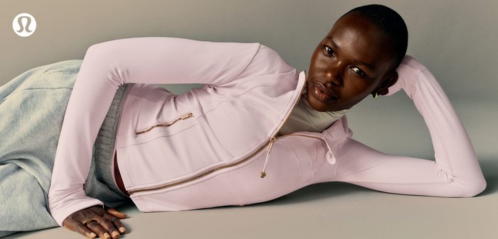 Persona sdraiata su un fianco che indossa una giacca con cerniera rosa chiaro e pantaloni della tuta grigi su uno sfondo neutro, con il logo Lululemon visibile.