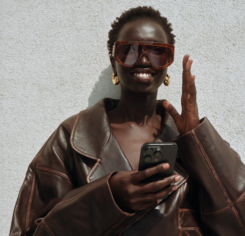Brown leather oversized jacket, beige fringed skirt, sheer tights, pointed stiletto heels, large rectangular sunglasses, sitting on grey stairs.
