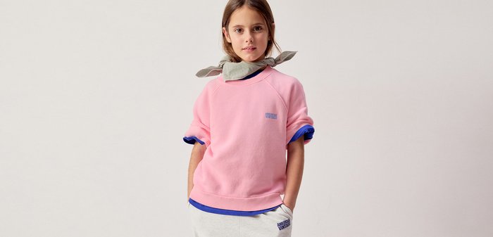 Ung pige står med hænderne i lommerne iført en pink sweatshirt over blå langærmede trøjer og lysegrå bukser med halstørklæde.