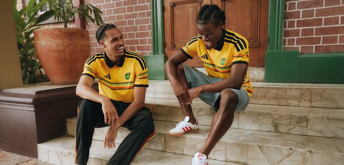 Δύο άντρες που φορούν κίτρινες φανέλες ποδοσφαίρου της Ιαμάικα κάθονται σε πέτρινα σκαλοπάτια, χαμογελούν και συνομιλούν κοντά σε ένα τούβλινο τοίχο και πράσινες πόρτες.
