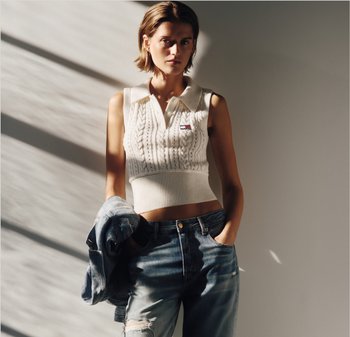 Vrouw die een mouwloze, wit gebreide cropped top en gescheurde spijkerbroek draagt, een spijkerjasje vasthoudend, staand tegen een grijze muur met schaduwlijnen.