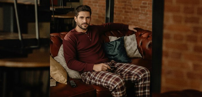 Donkerrood shirt met lange mouwen, geruite broek in tinten bruin en crème, zittend op een bruine leren bank, een afstandsbediening vasthoudend.