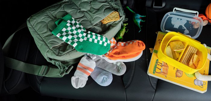Sac à dos matelassé vert avec des chaussettes colorées pour enfants, des dinosaures en jouet, une boîte à lunch jaune avec des crackers, et un livre sur un siège de voiture.