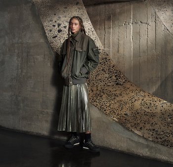 Giacca bomber verde oliva con polsini a costine, gonna midi plissettata metallica e sneakers nere chunky con accenti bianchi, posizionate su uno sfondo testurizzato.