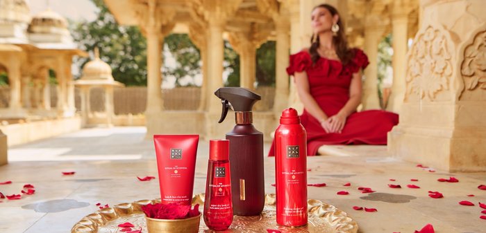 Produits de soin de la peau Red Ritual of Ayurveda disposés sur un plateau doré avec des pétales de rose, femme en robe rouge assise floue en arrière-plan.