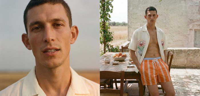 Jeune homme aux cheveux courts et foncés, portant une chemise crème ouverte et un short rayé orange, debout près d'une table en bois avec des fruits en plein air.