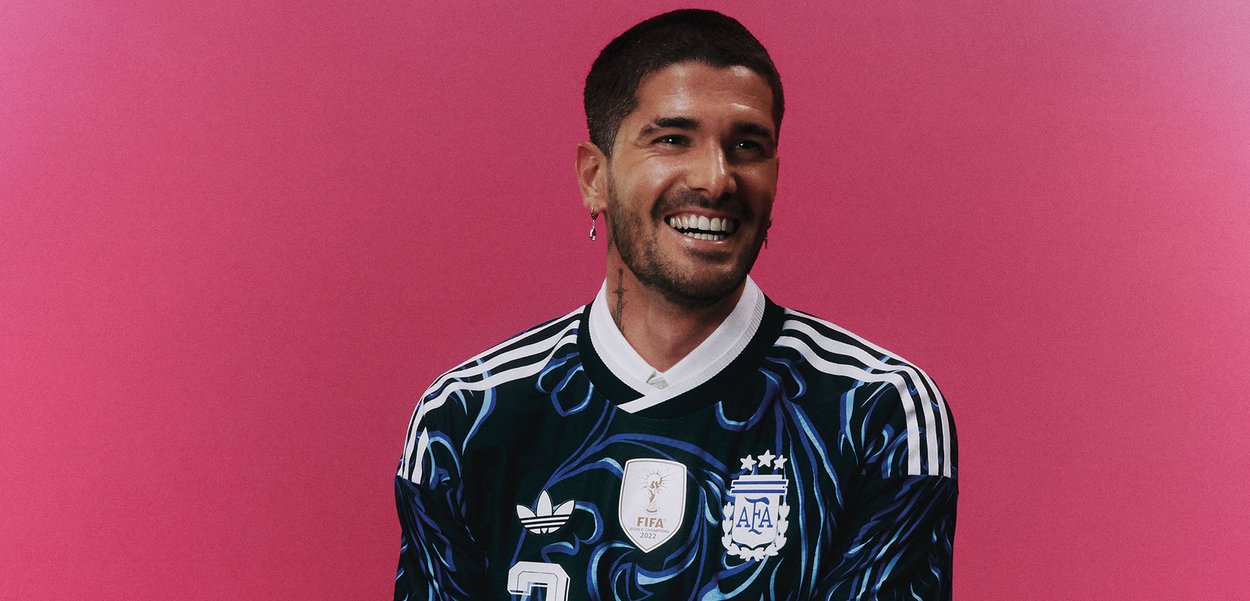 Homme souriant aux cheveux courts et foncés porte un maillot de football sombre de l'Argentine avec des bandes Adidas et un écusson FIFA, debout devant un fond rose.