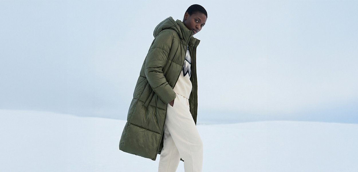 Person wearing a green hooded padded coat, white patterned jumper, and white trousers, standing in a snowy outdoor landscape.