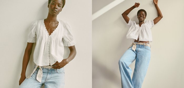 Woman wearing a white blouse and light blue jeans with a patterned belt poses standing and with arms raised against a plain wall.