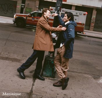 Two men stand near a street pole. One wears a brown coat and black shoes; the other wears a blue jacket and brown pants with dark boots.