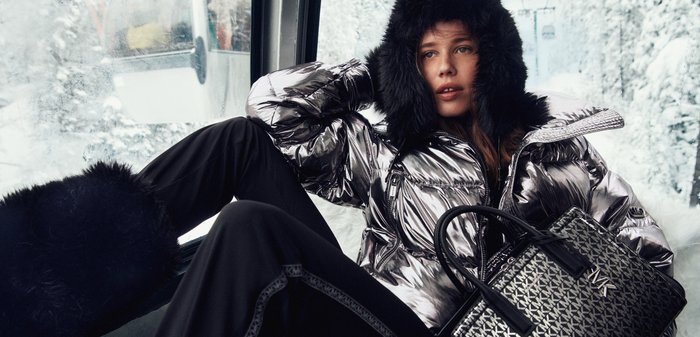 Glänzende silberne Steppjacke mit kapuzenbesatz mit Pelz, kombiniert mit schwarzen Hosen und einer gemusterten schwarzen Handtasche mit Logoakzent.