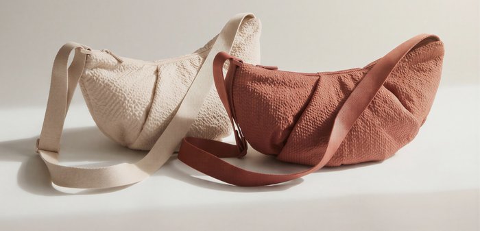 Two soft-textured crescent-shaped shoulder bags with adjustable straps, one beige and one rust-colored, placed on a light surface.