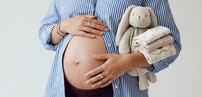 Έγκυος άτομο με ριγέ πουκάμισο που κρατά την κοιλιά με το ένα χέρι και ένα απαλό παιχνίδι μαζί με διπλωμένα βρεφικά ρούχα στο άλλο.