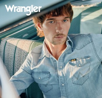 Jeune homme aux cheveux châtain clair portant une chemise en denim clair, assis dans une voiture vintage avec des sièges à motifs. Logo Wrangler en haut à gauche.