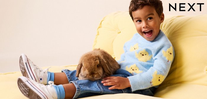 Fiatal gyermek kék csibemintás pulóverben, sárga kanapén ülve, mosolyogva, és barna nyulat tartva az ölében.