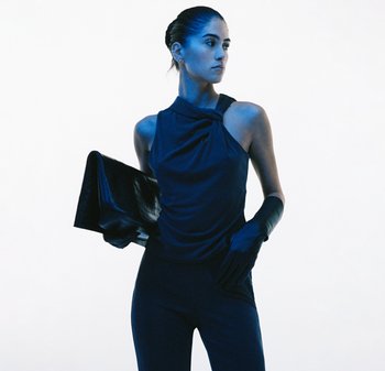 Marineblå ærmeløs top med draperet halsudskæring, kombineret med tætsiddende mørke bukser og sort clutch. Modellen bærer lange sorte handsker. Baggrunden er hvid.