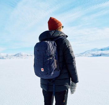 Navyblauer Rucksack aus strapazierfähigem Stoff mit einem vorderen Bungee-Kordeldesign, getragen auf dem Rücken einer Person in einer Steppjacke und einer Mütze.