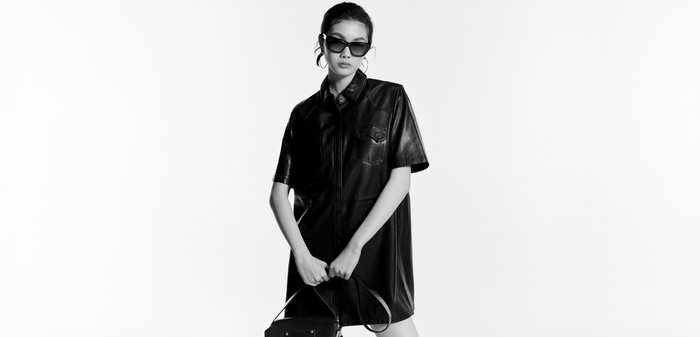 Femme portant une robe-chemise en cuir noire, des lunettes de soleil et des boucles d'oreilles créoles, tenant un petit sac à main noir à deux mains.