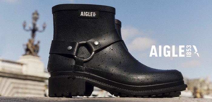 Bottines noires imperméables avec une surface texturée, un accent en anneau argenté et une semelle en caoutchouc épaisse. Présente une étiquette de marque près du haut.