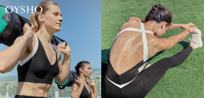 Two women in black sports bras carry weighted bags on their shoulders, exercising outdoors under a clear blue sky.