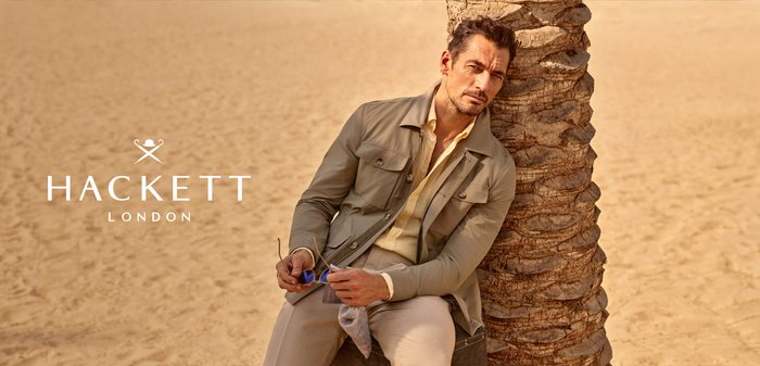 Hombre con chaqueta beige y pantalones claros sentado junto a una palmera en un desierto arenoso, sosteniendo gafas de sol, con el logo de Hackett London a la izquierda.