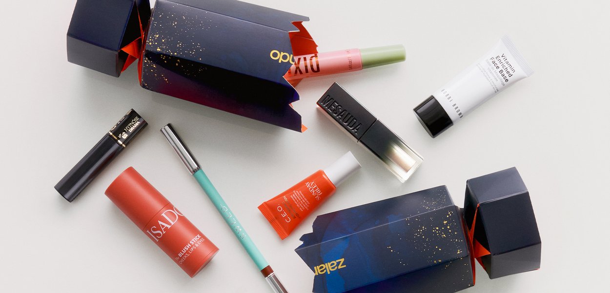 Deux boîtes festives ouvertes de couleur bleu marine avec des éclats dorés contenant divers cosmétiques : blush pour les lèvres et les joues, mascara, primer pour le visage, eyeliner et baume à lèvres.