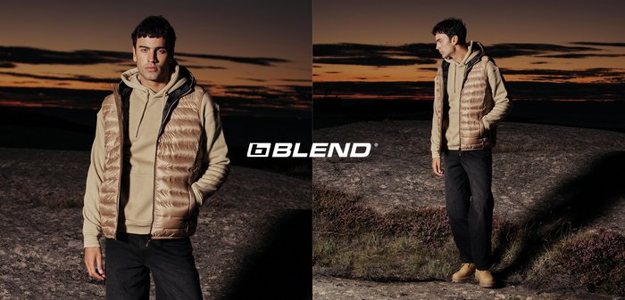 Junger Mann in beiger Kapuzenjacke, beige gesteppter Weste, schwarzer Hose und Stiefeln steht bei Sonnenuntergang auf felsigem Gelände, mit Blend-Logo zwischen zwei Posen.