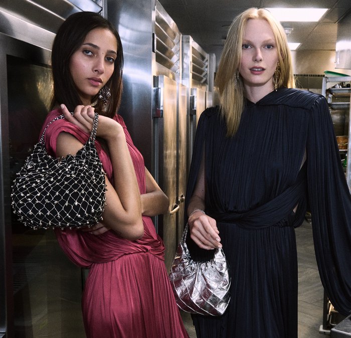 Dwie kobiety pozują w pomieszczeniu. Jedna trzyma czarną, ozdobioną ćwiekami torebkę; druga ma metaliczną, srebrną torbę. Obie noszą zwiewne, eleganckie sukienki.
